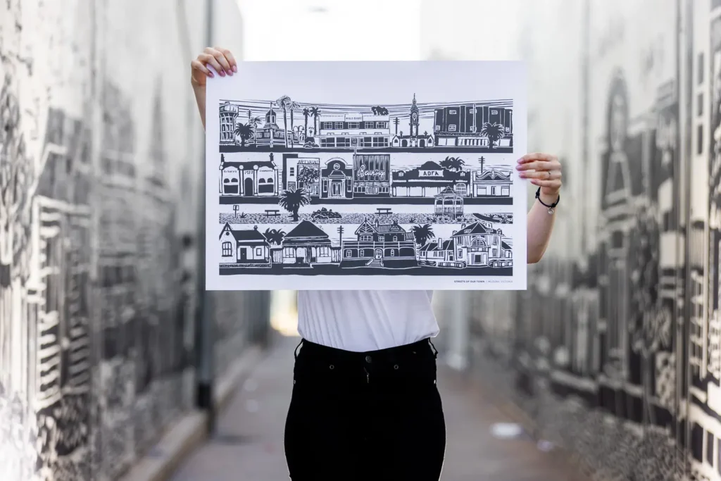 A person holding up the Streets of our Town poster against the mural artwork behind them