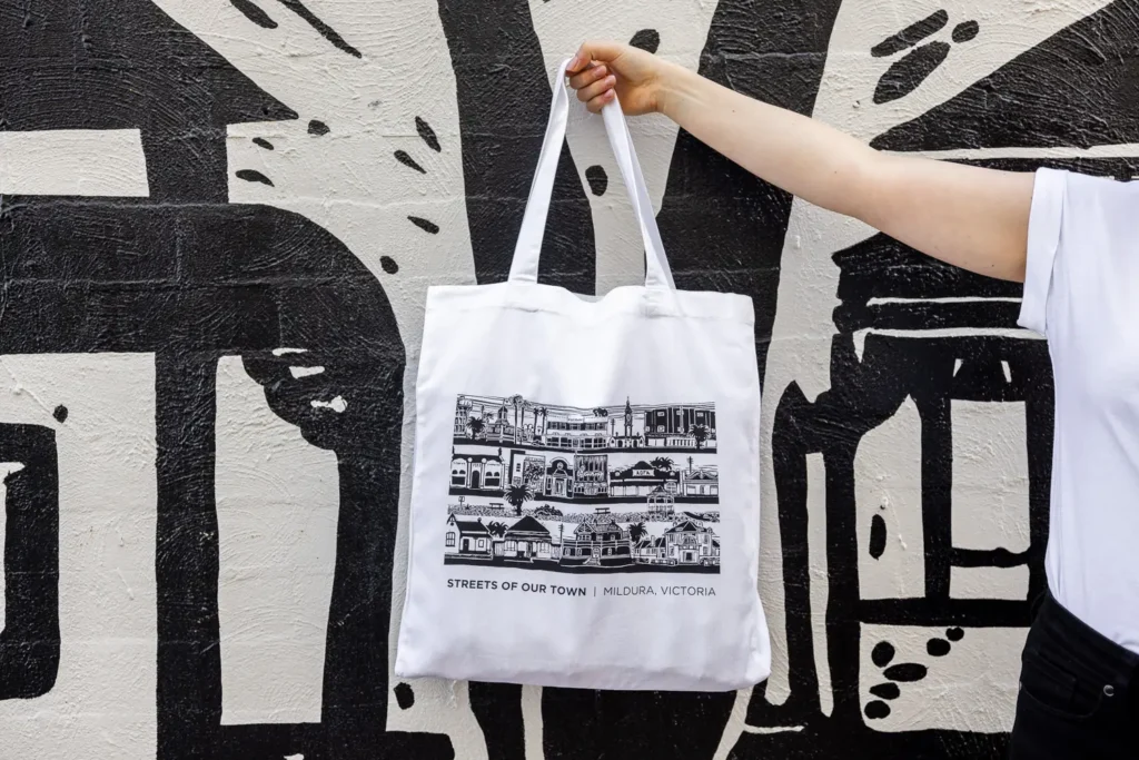 A person holding up the Streets of our Town tote bag against the mural artwork behind them